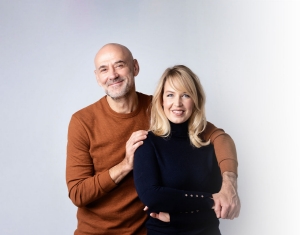 Mature couple stands close together.  The man has his arm over the woman's shoulders.