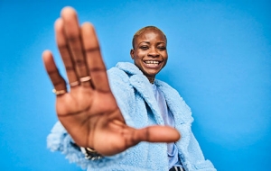 A young woman of color smiles and waves to the camera.