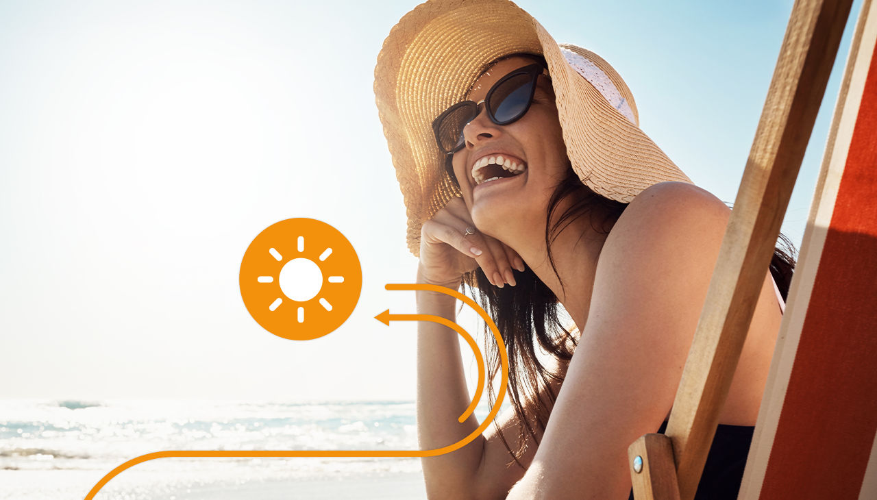 A smiling woman holds on to her hat at the beach.